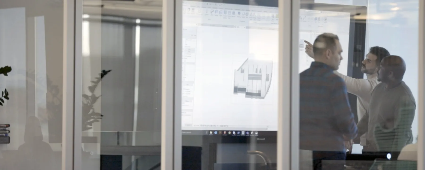 Three engineers behind a glass partition reviewing a 3D model on screen, one pointing at the display
