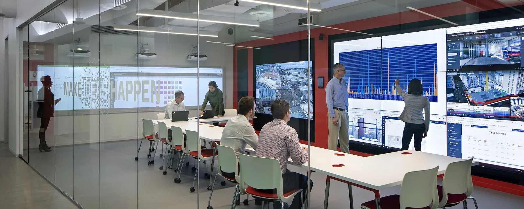 Team seated around a long table in a glass-walled room facing a wall of project dashboards