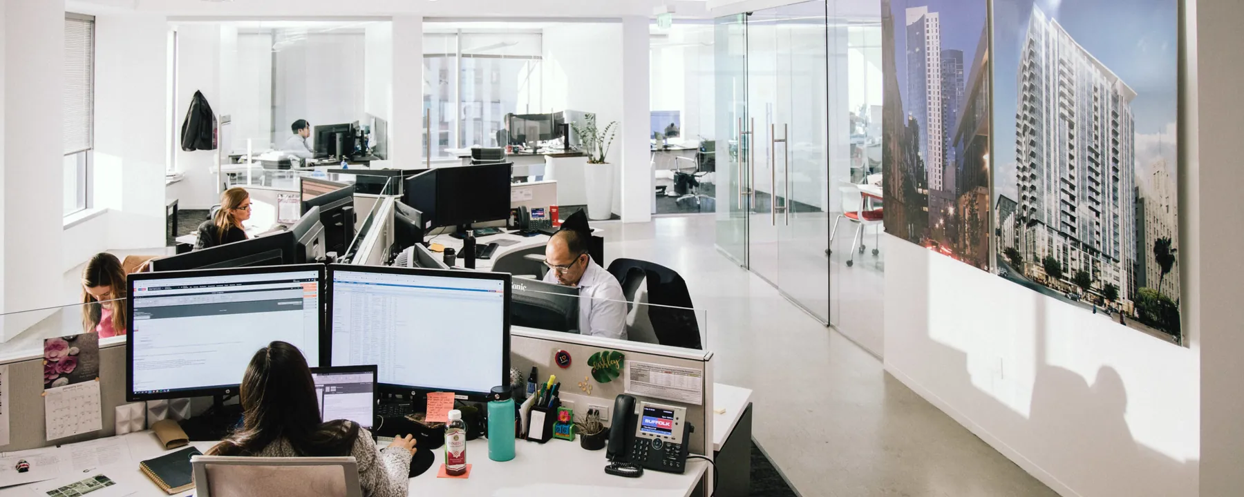 Open-plan project office with staff at dual-monitor workstations and a large building rendering on the wall