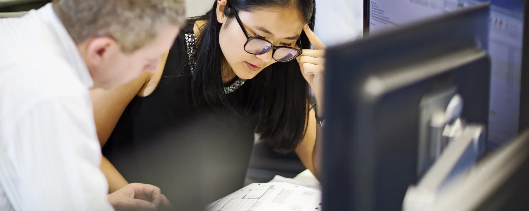 Two colleagues leaning over a desk studying a monitor together, printed documents in hand