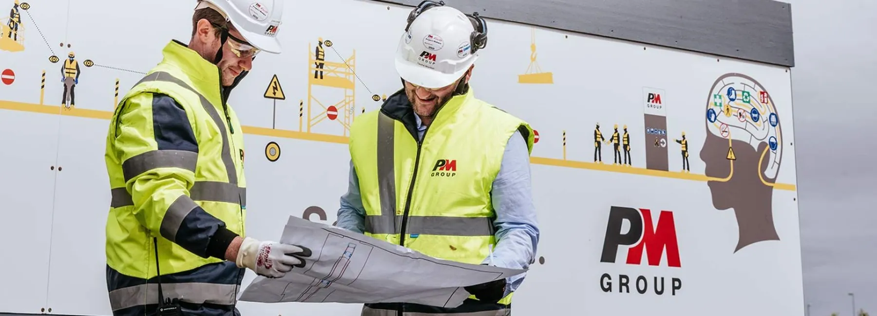 Two workers in high-visibility jackets and hard hats reviewing a large drawing on a construction site