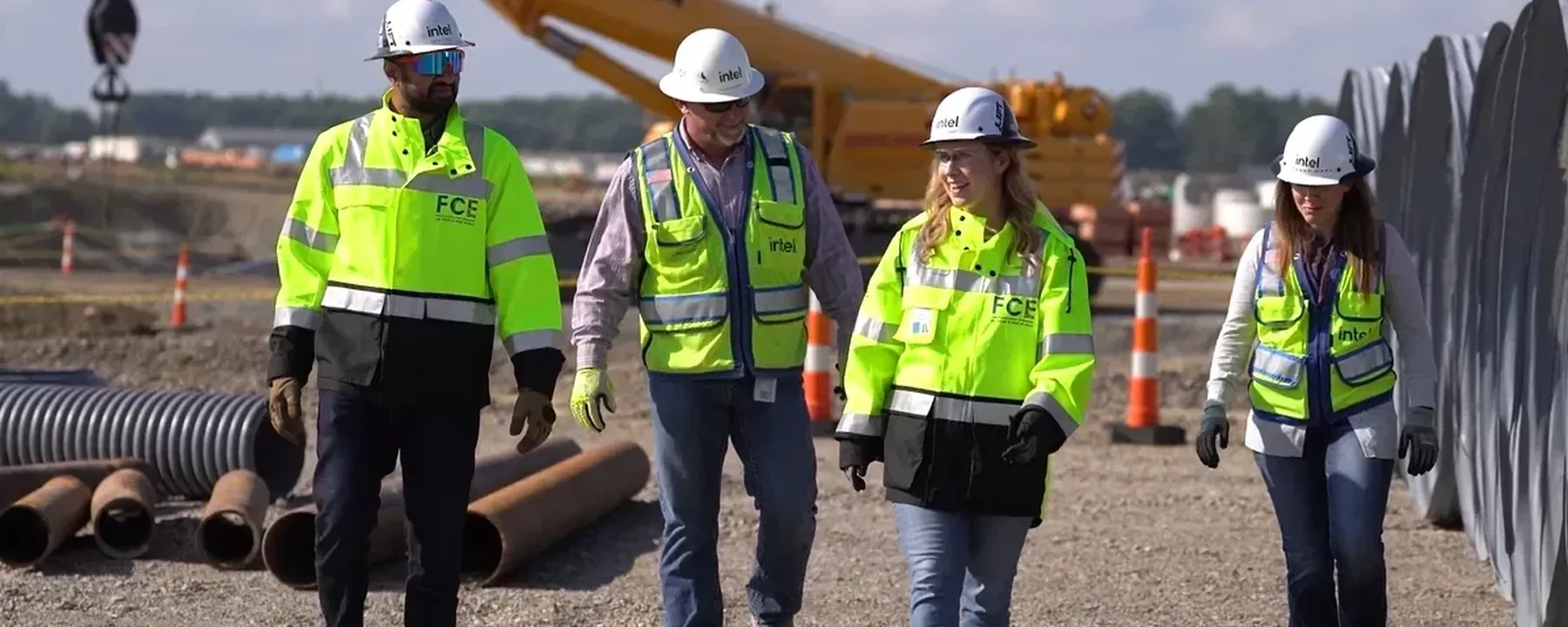 Four workers in high-visibility gear walking through a laydown yard past stacked pipe, a crawler crane behind them