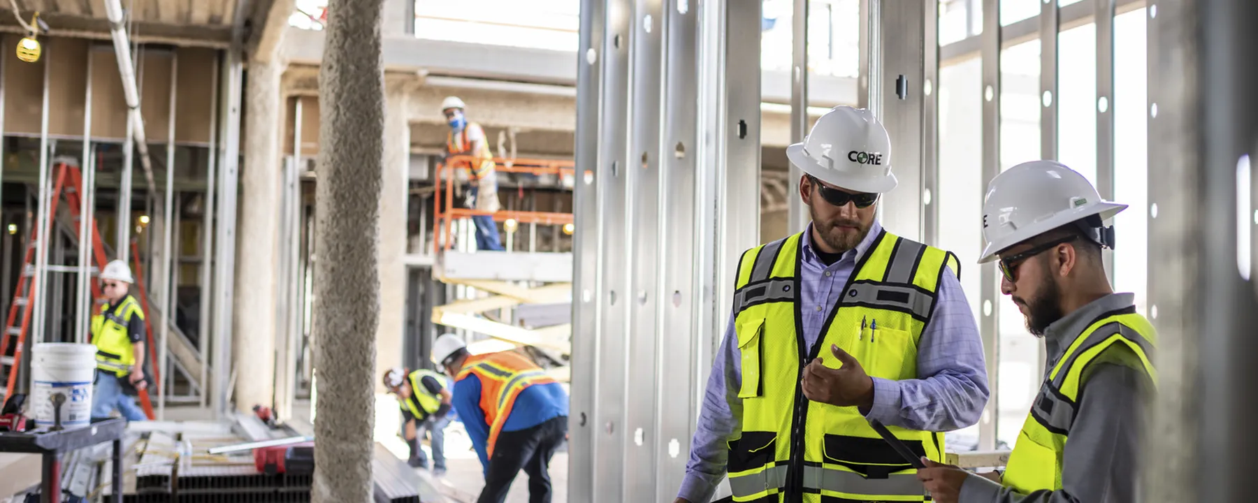 Two workers in hard hats and high-visibility vests reviewing a document inside a building under construction, metal stud framing around them