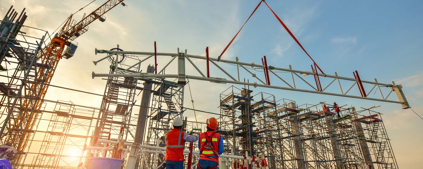 Two workers guiding a steel truss lowered by a tower crane over a scaffolded structure at sunrise